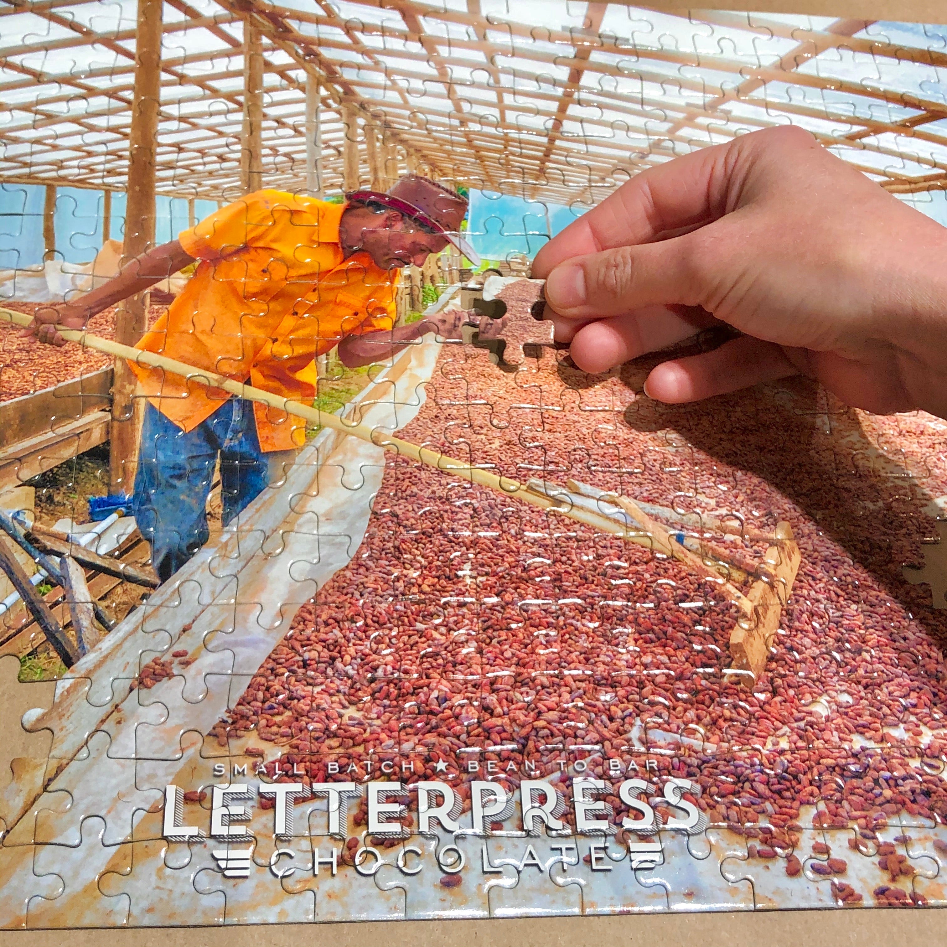 Detail picture of a hand placing a puzzle piece of a Puzzle showing a cacao farmer inspecting cacao beans in a solar dryer